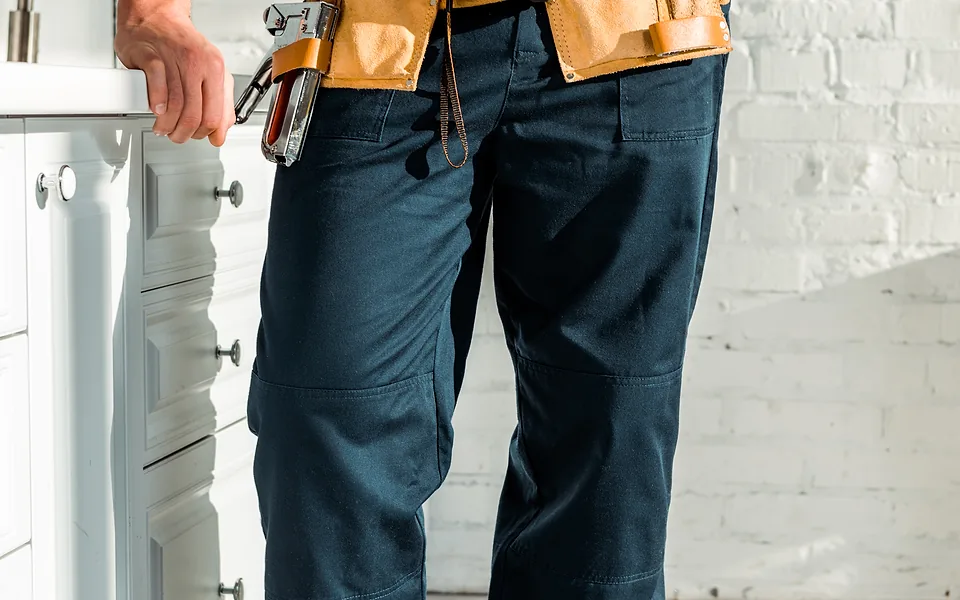Worker in navy work pants and a leather tool belt standing beside white kitchen cabinets, shown waist down