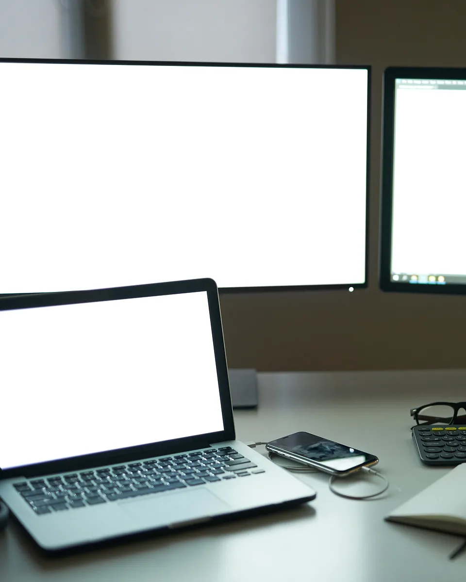 Laptop and two desktop monitors showing blank white screens on a desk with a phone and glasses