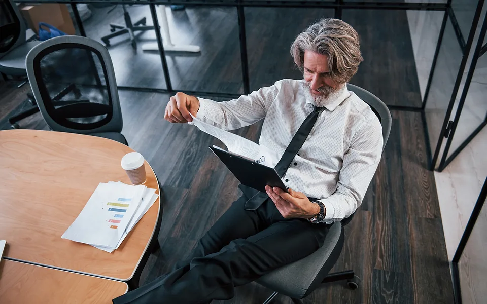 Grey-haired man in a shirt and tie leaning back in an office chair, leafing through a folder of papers