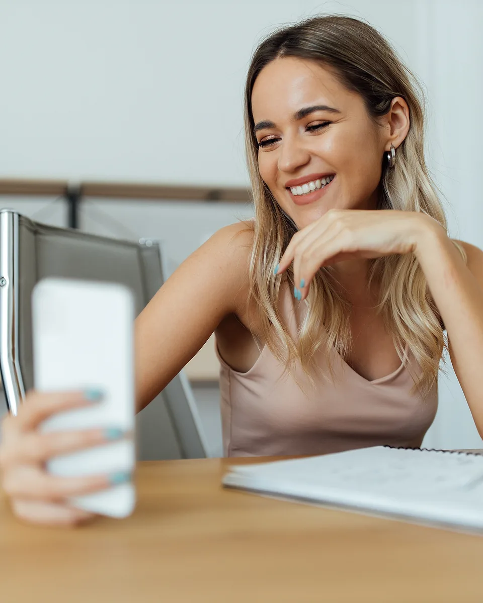 Smiling woman with long hair looking at the phone she is holding up, seated at a desk with a notebook