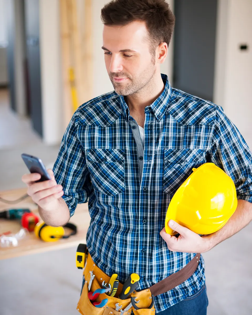 Tradie in a blue checked shirt and tool belt reading his phone on site, a yellow hard hat under his arm
