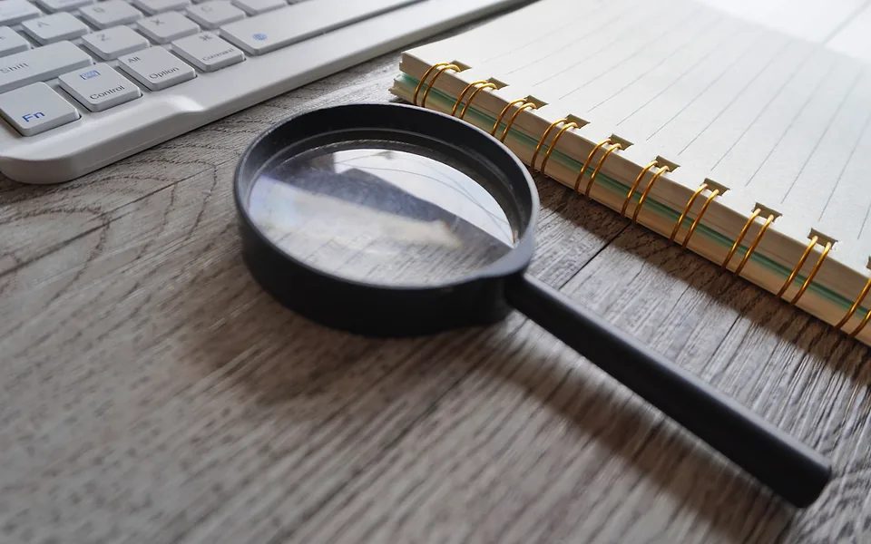 Black magnifying glass on a timber desk between a white keyboard and a spiral-bound lined notebook