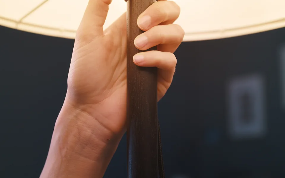 Hand gripping the timber stem of a lamp beneath its glowing shade in a dimly lit room