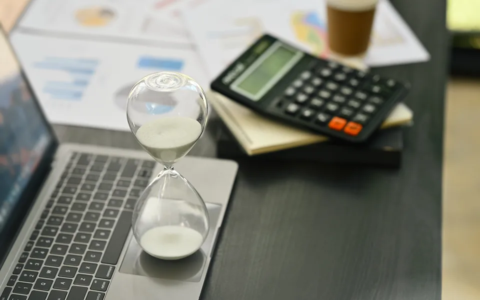 Sand running through an hourglass set on a laptop, beside a calculator and printed charts