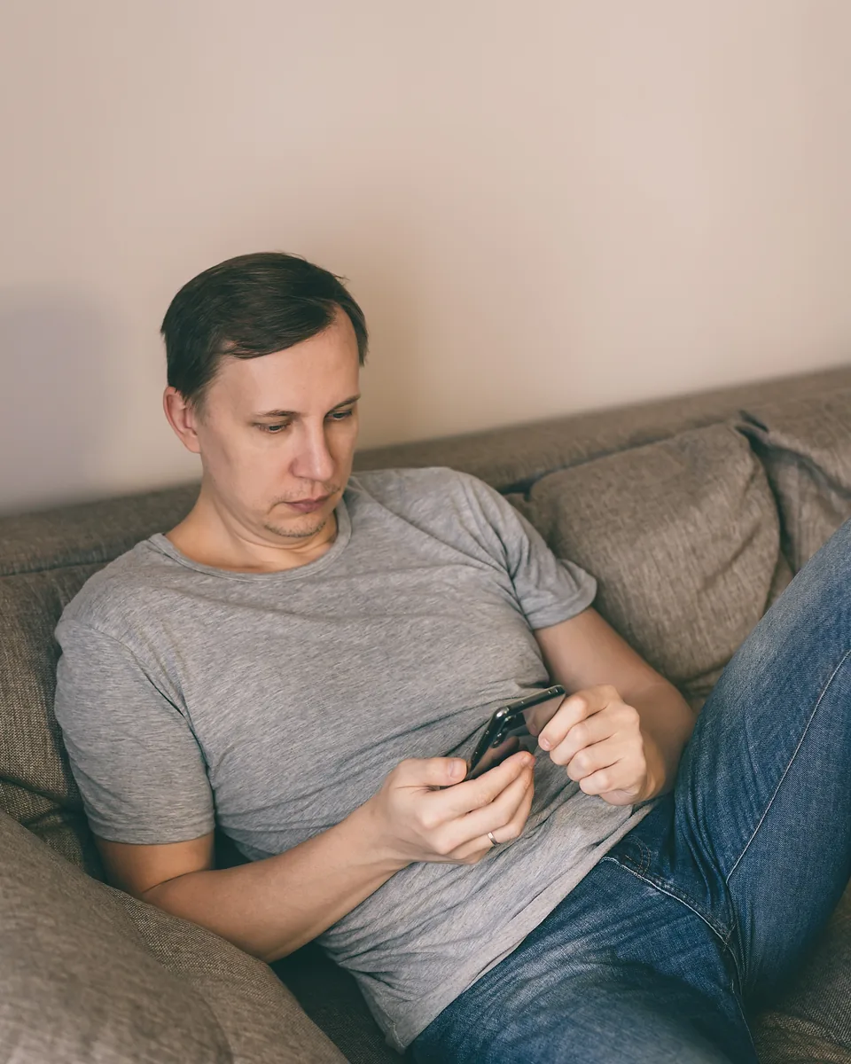 Man in a grey t-shirt and jeans lying back on a couch and scrolling on his phone with both hands