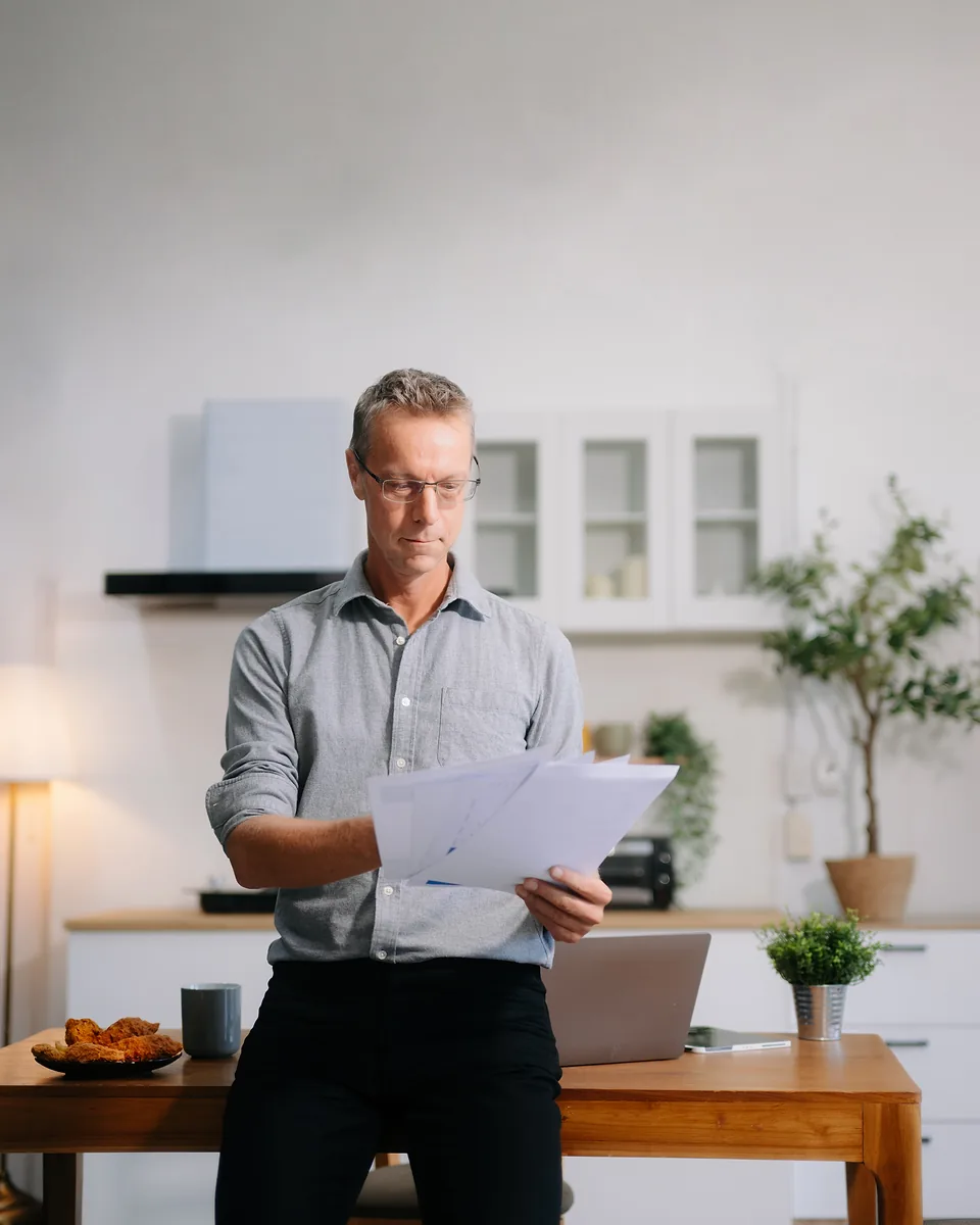 Grey-haired man in glasses leaning on a kitchen table and reading through a handful of printed pages