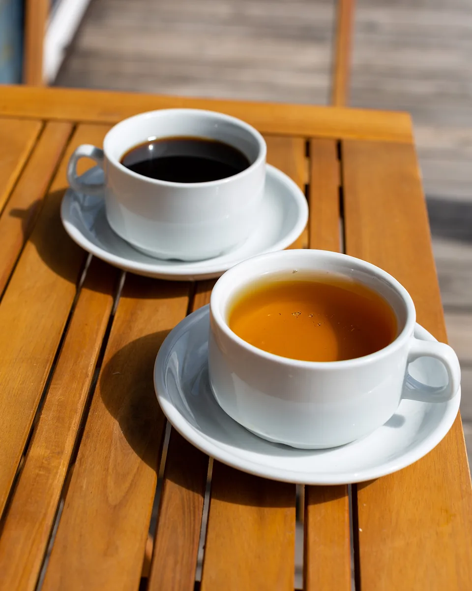 Two white cups on saucers, one of black coffee and one of tea, on a slatted timber outdoor table