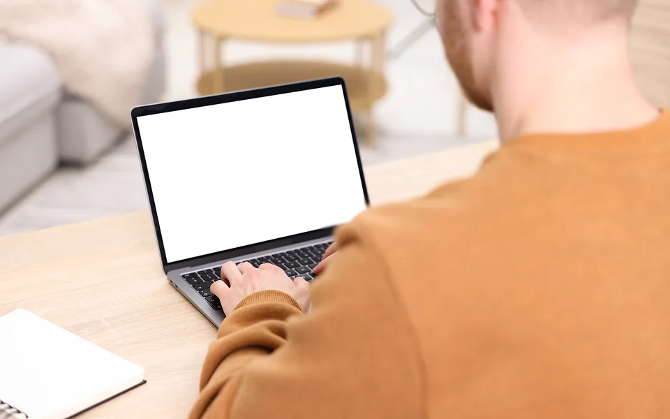 Over-the-shoulder view of a man in an orange jumper typing on a laptop with a blank white screen