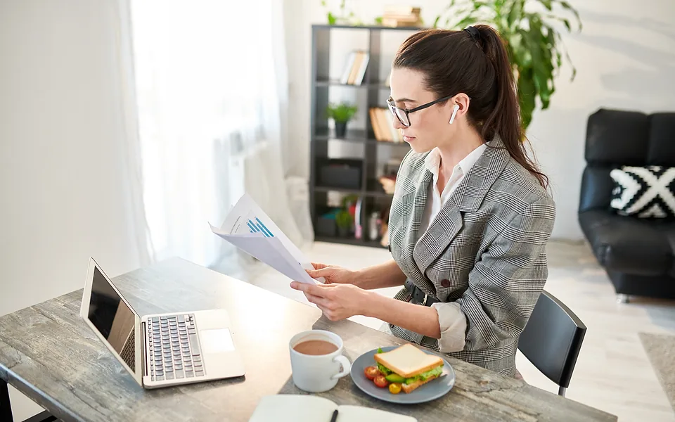 Woman in glasses and a grey blazer reading a printed chart at a table with a laptop, tea and a sandwich