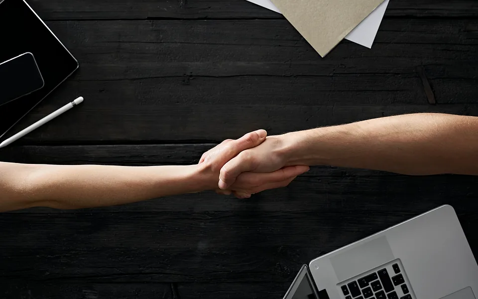 Top-down view of two people shaking hands across a black timber desk with a laptop, tablet and stylus