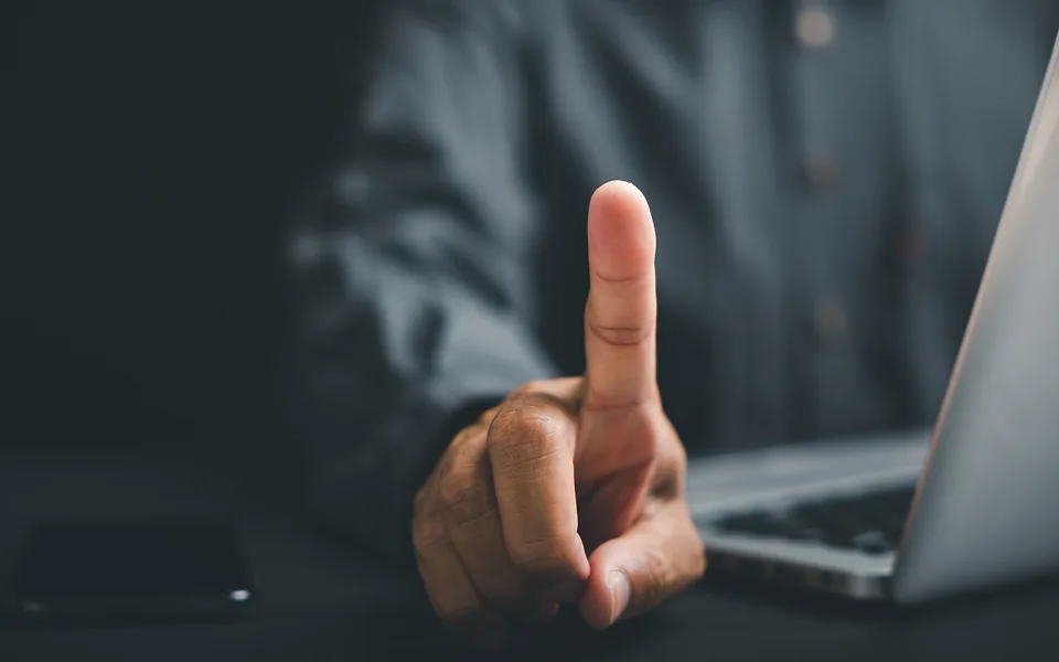 Man in a dark shirt holding up his index finger in front of an open laptop in low light