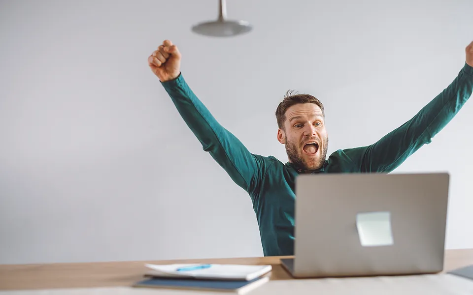 Bearded man in a green jumper cheering with both fists in the air in front of his laptop