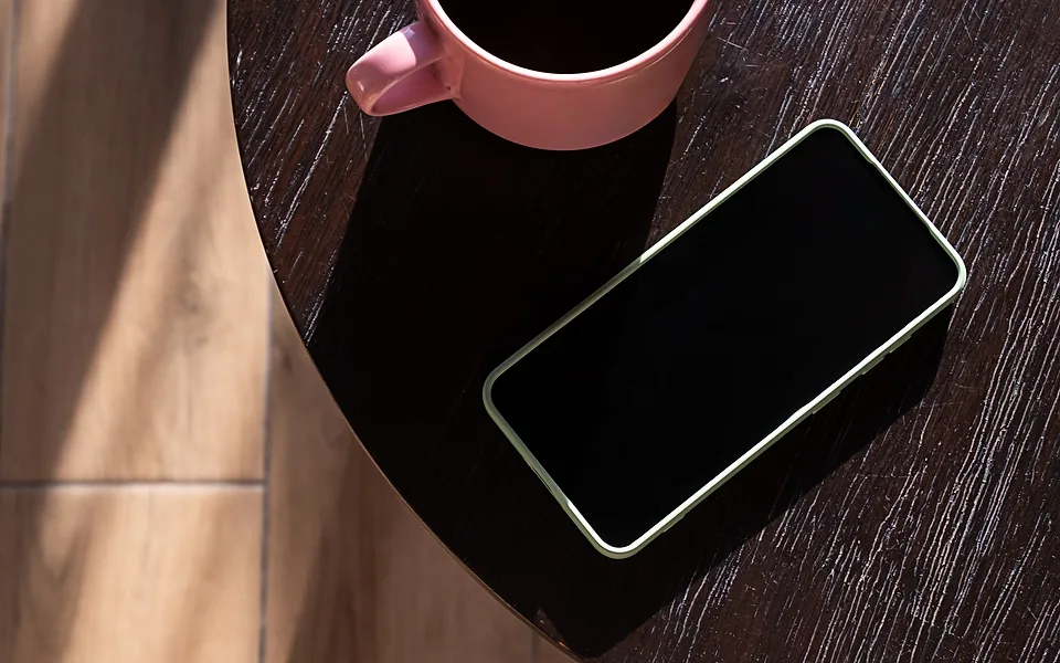 Dark-screened phone lying on a round dark timber table next to a pink coffee cup, seen from above