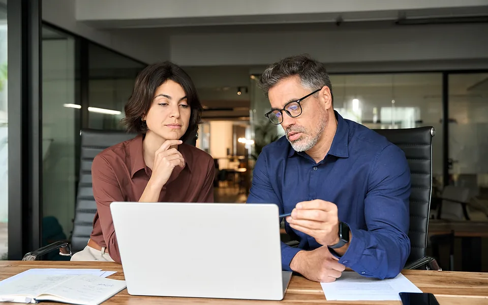 Woman and man in glasses sitting together at a desk and looking over a laptop in a glass-walled office