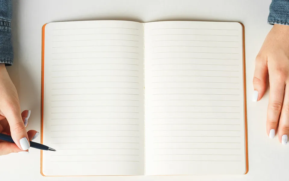 Open lined notebook with blank pages on a white desk, one hand holding a pen at its edge