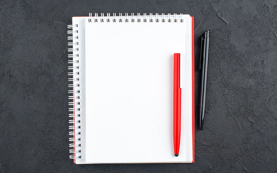 Blank spiral notepad with a red pen resting on it and a black pen beside it on a dark slate surface