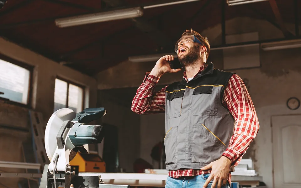 Tradie in a checked shirt and work vest laughing during a phone call in his timber workshop