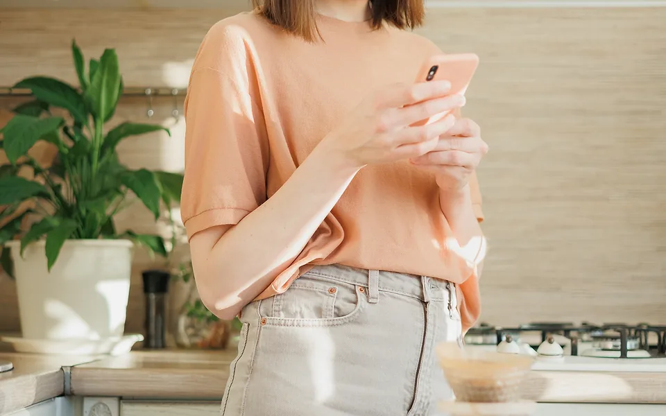 Woman in a peach top holding a phone in both hands in a kitchen with a large pot plant behind her