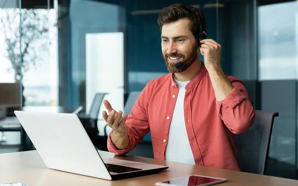 Smiling bearded man in a coral shirt wearing a headset and gesturing as he talks at a laptop