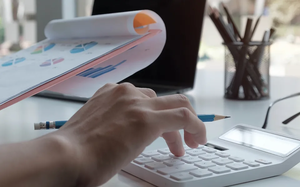 Hand tapping a white calculator next to printed pages of pie and bar charts on a desk