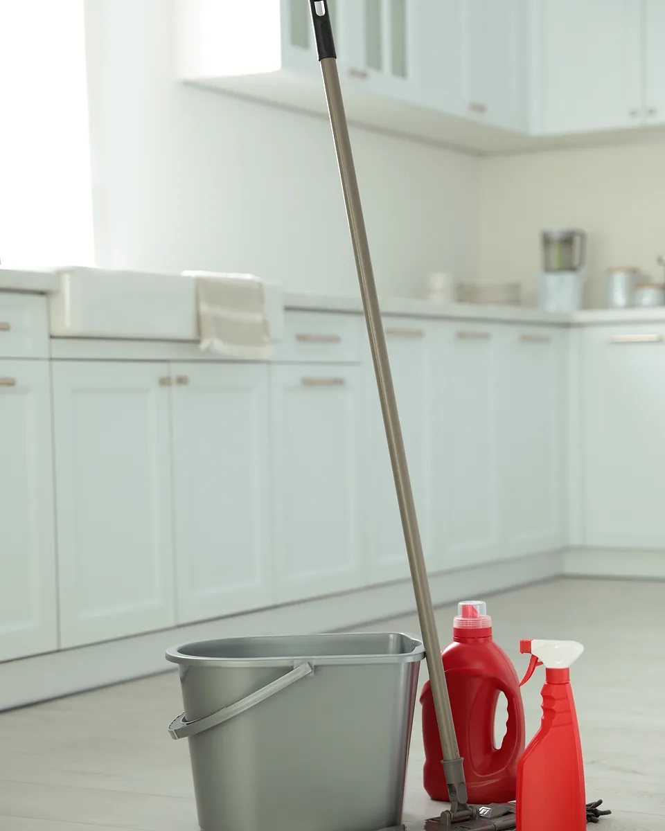 A cleaner's mop, grey bucket and red spray bottles on the floor of a spotless white kitchen