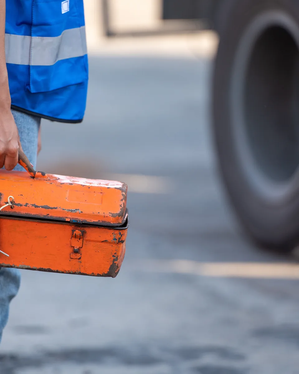 Tradie in a blue hi-vis vest and jeans carrying a worn orange metal toolbox past a large tyre