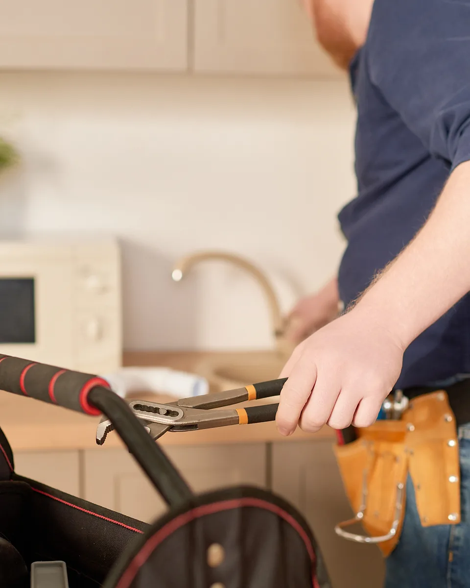 Close-up of a tradie's hand holding multigrip pliers over an open tool bag in a home kitchen