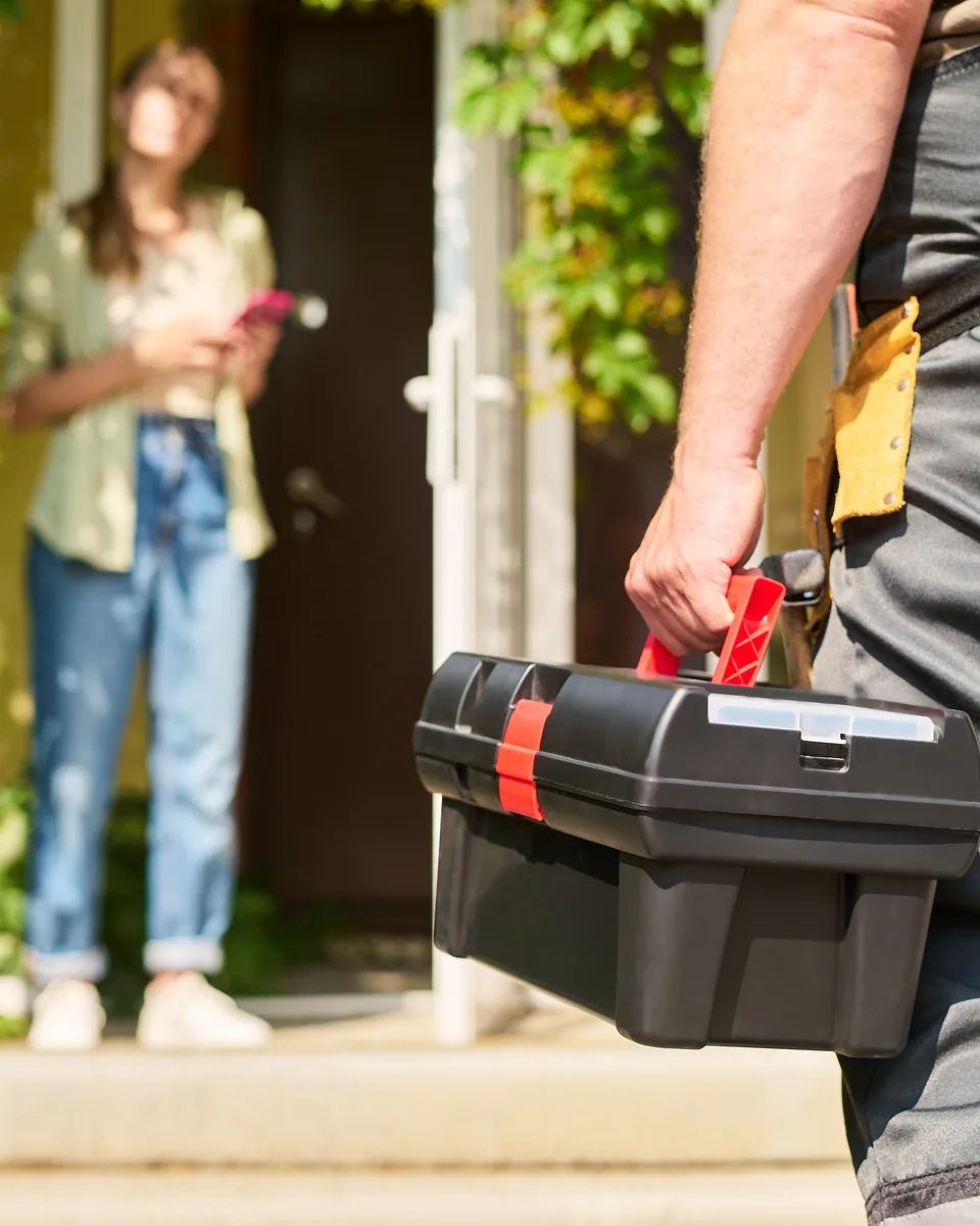 Tradie with a tool belt and black toolbox walking up to a front door where a homeowner stands waiting