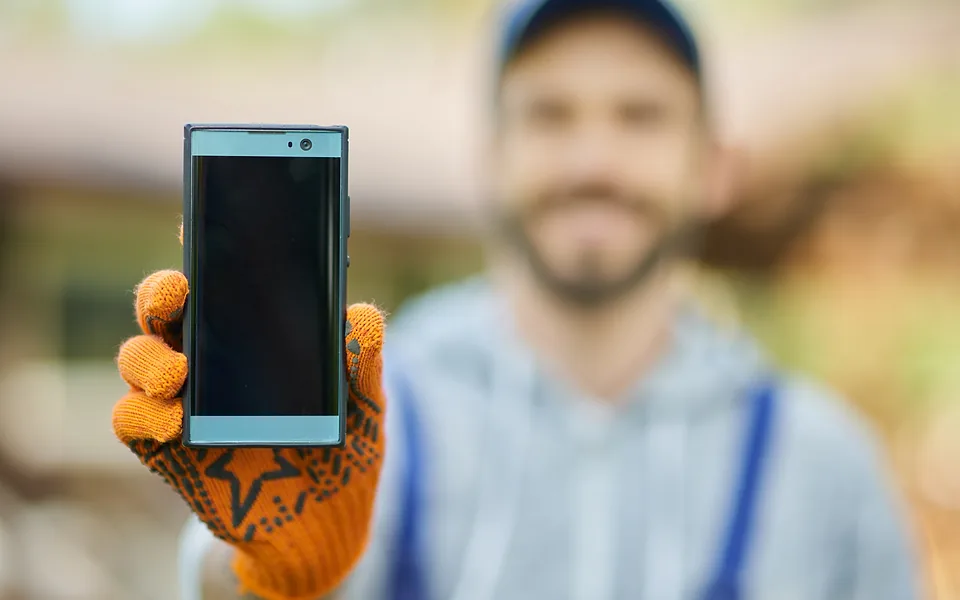 Tradie in a cap and work gloves holding a dark-screened phone towards the camera, his face out of focus