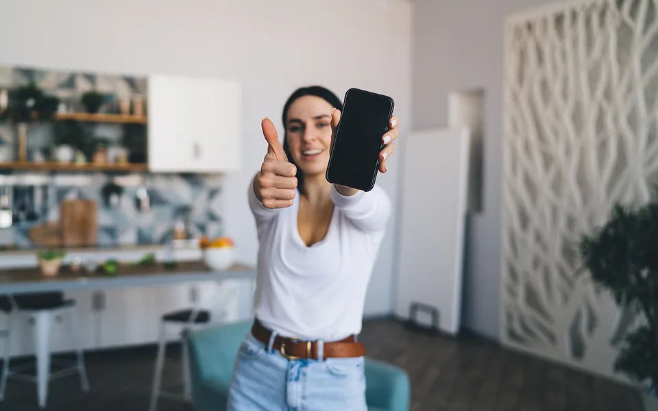 Smiling woman in a white top giving a thumbs up and holding out a dark-screened phone in a kitchen