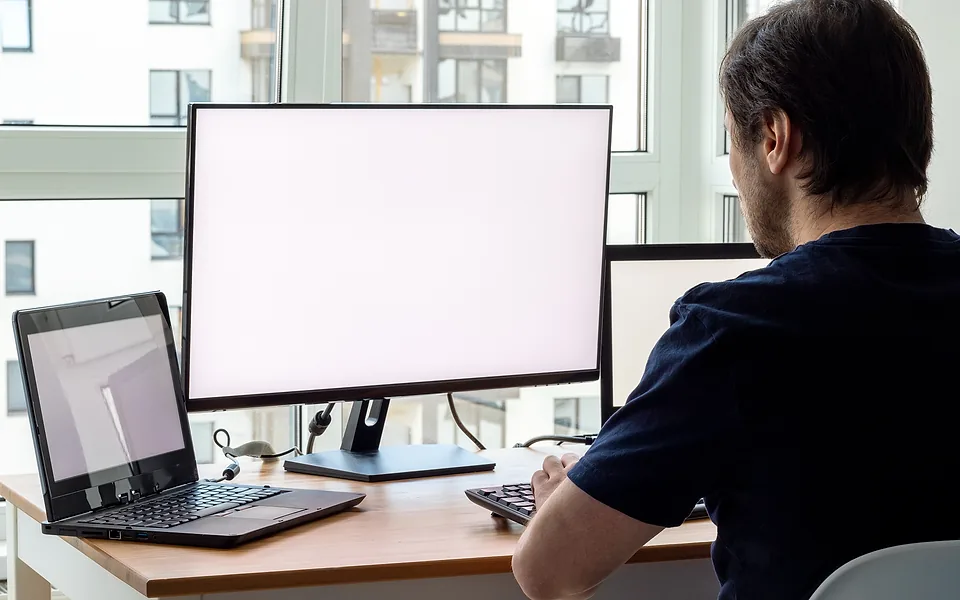 Desk with a blank widescreen monitor and a laptop, viewed over the shoulder of a man in a dark t-shirt