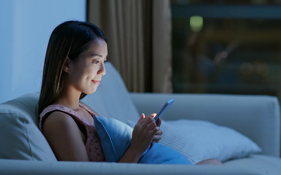Woman lying on a sofa at night, her face lit by the phone she is scrolling through