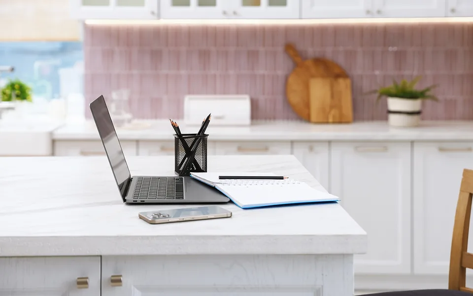 Open laptop beside a phone, notebook and pen holder on a white kitchen island with pink tiles behind
