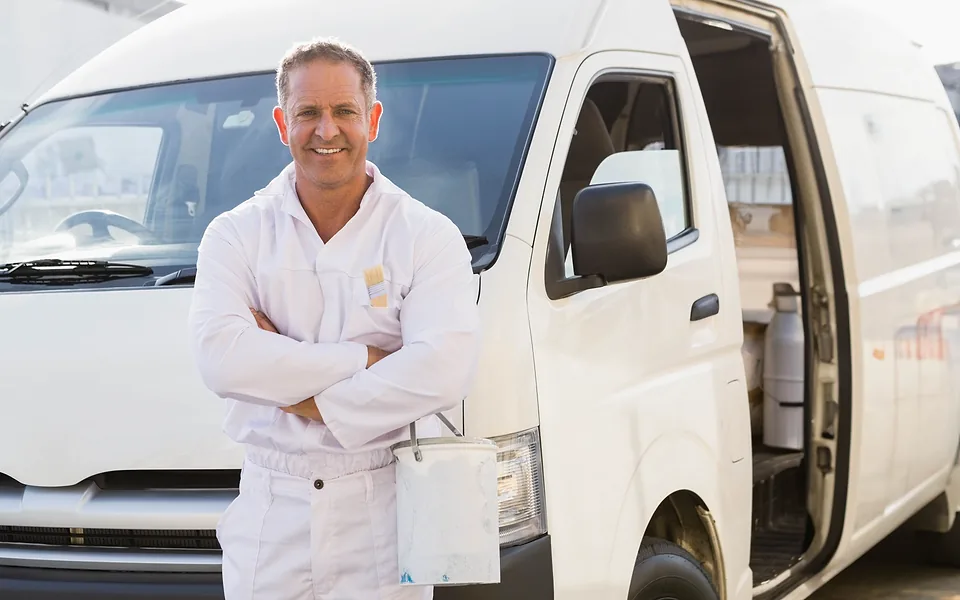 Tradie in white painter overalls standing with his arms crossed in front of a white work van