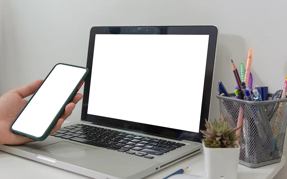 Hand holding a phone beside an open laptop on a white desk, both showing blank white screens