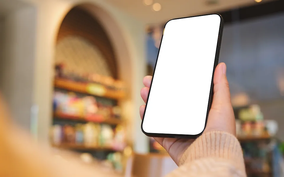Hand in a knitted sleeve holding up a phone with a blank white screen, with blurred shelves behind