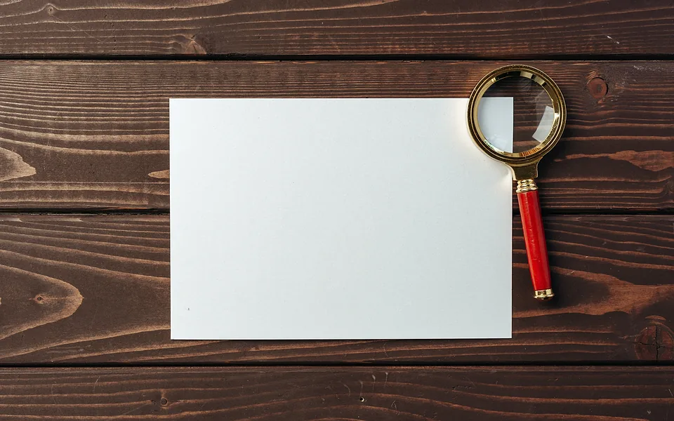 Brass magnifying glass with a red handle lying on a blank sheet of white paper on dark timber boards