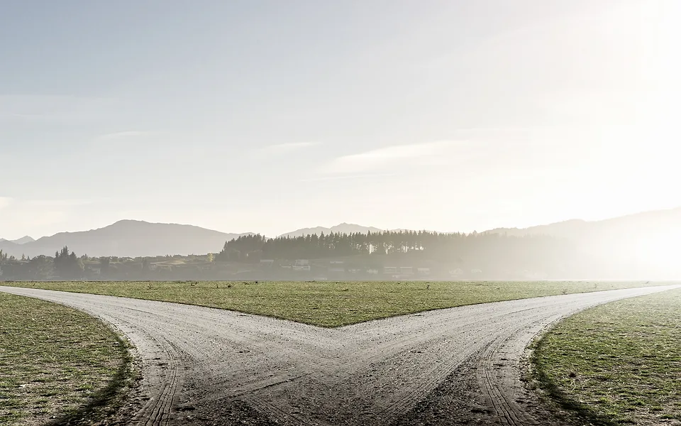 Gravel road forking into two tracks across a flat grassy field, with misty hills in the distance