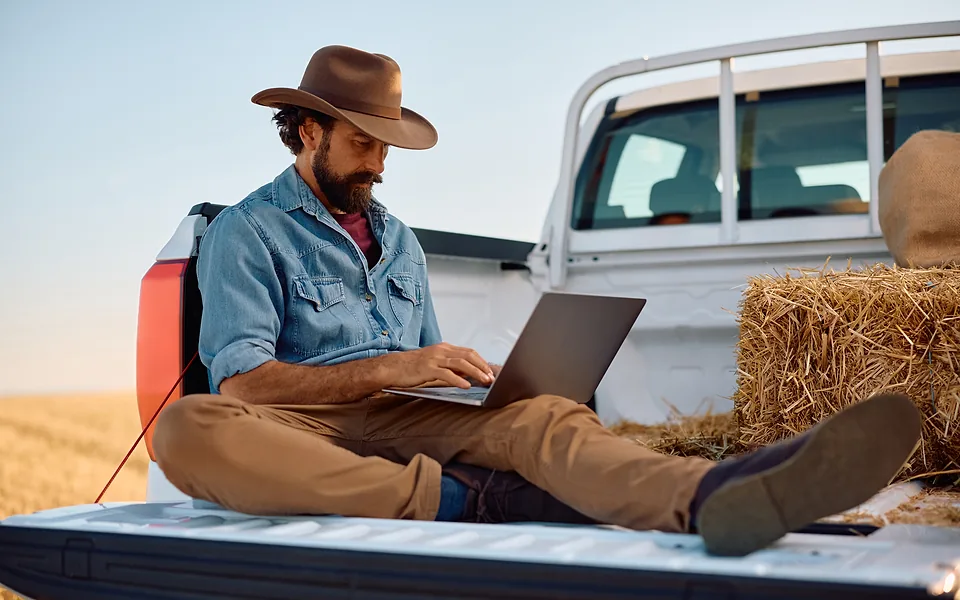 Bearded man in a wide-brimmed hat sitting on a ute tray with a laptop, hay bales stacked beside him