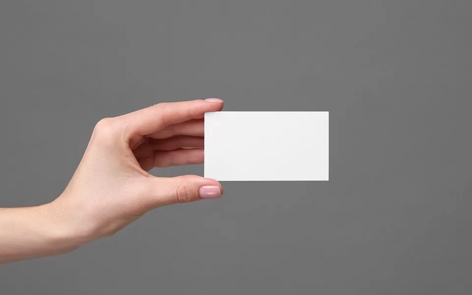 Hand holding out a blank white business card between finger and thumb against a plain grey background