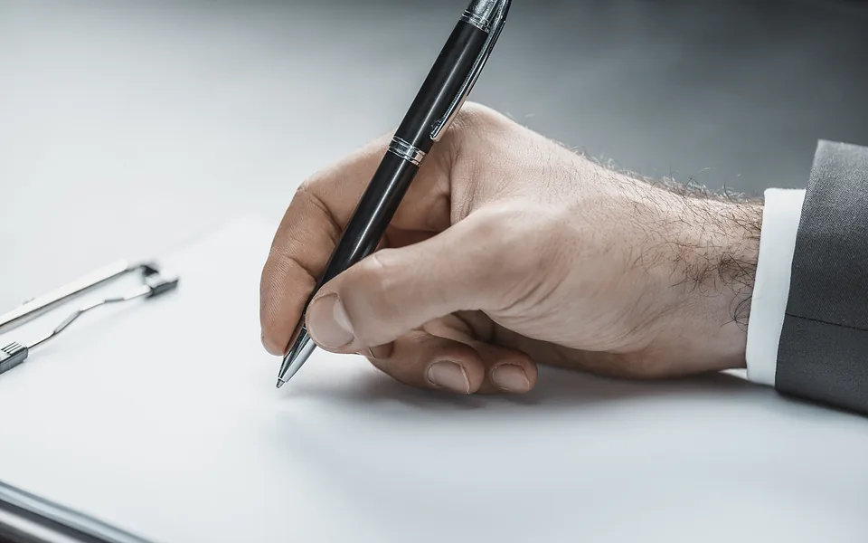 Hand in a dark suit sleeve signing a sheet of paper on a clipboard with a black ballpoint pen