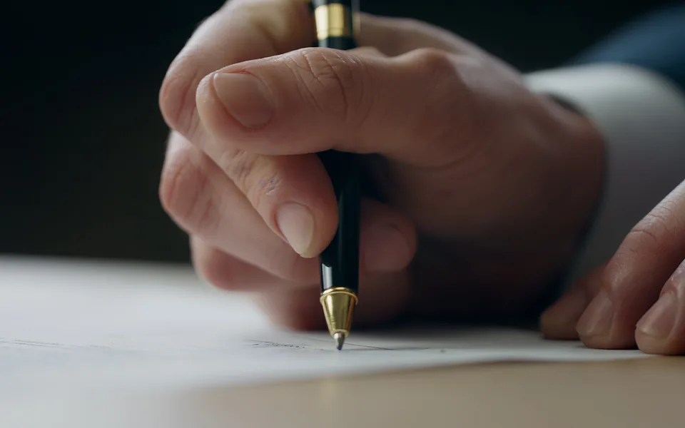 Close-up of a hand signing a document with a black and gold pen while the other hand steadies the page