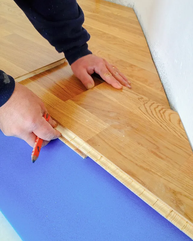 Flooring tradie's hands laying timber boards onto bright blue underlay beside a white wall