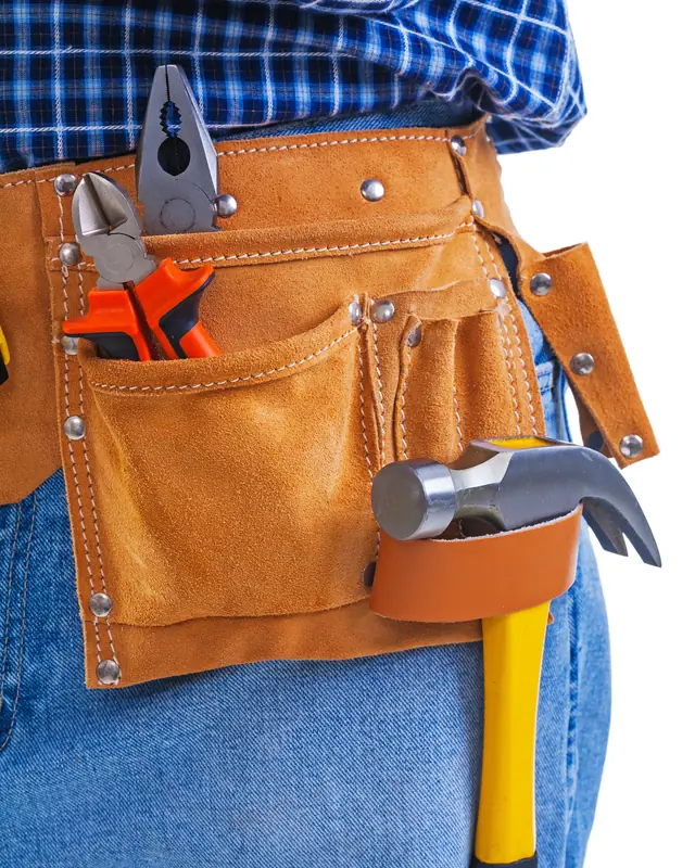 Pliers, side cutters and a claw hammer sit in the tan leather tool belt of a handyman in jeans