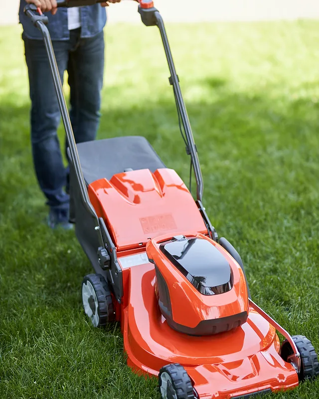 Worker on a lawn mowing job, in jeans and a checked shirt, pushing an orange mower over thick grass
