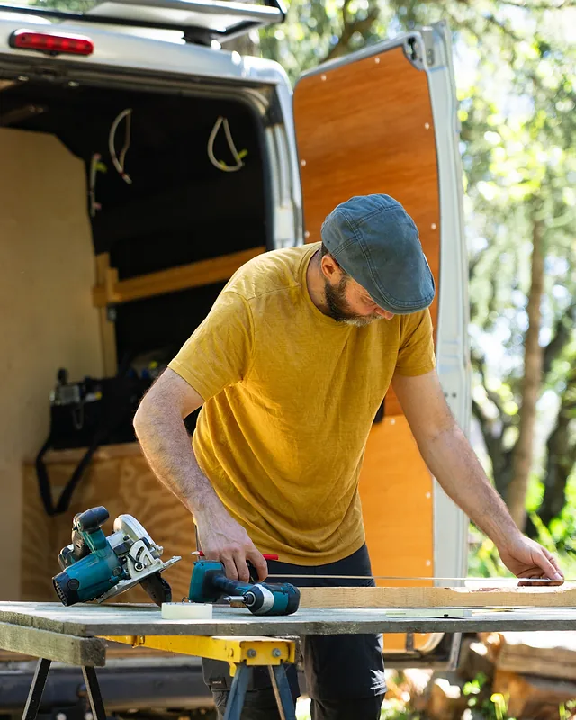 An open work van stands behind a tradesman in a flat cap as he lines up timber on a portable bench