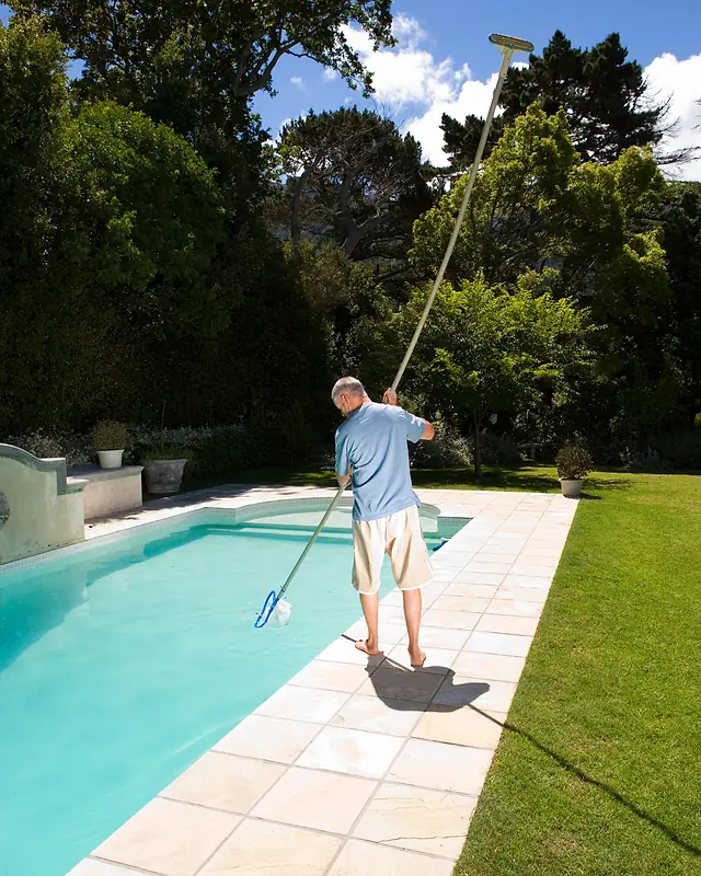 Pool and spa upkeep in a backyard, with a man in shorts brushing a turquoise pool beside lawn and tall trees