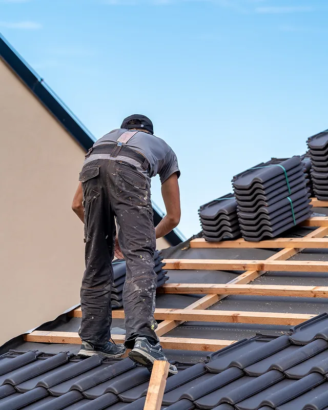 Roofer standing on timber battens beside stacks of dark roof tiles, working under a clear blue sky