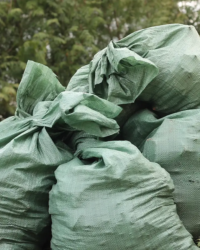 Tied green woven rubbish bags piled in front of trees, a typical load for a local rubbish removal business