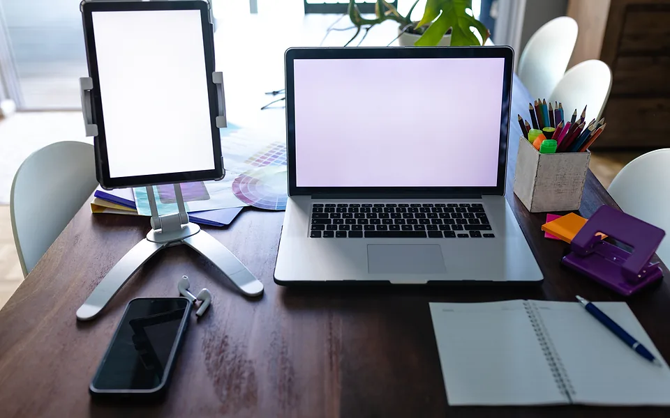 Tablet on a stand and a laptop, both with blank screens, on a dark timber table with a phone and notebook